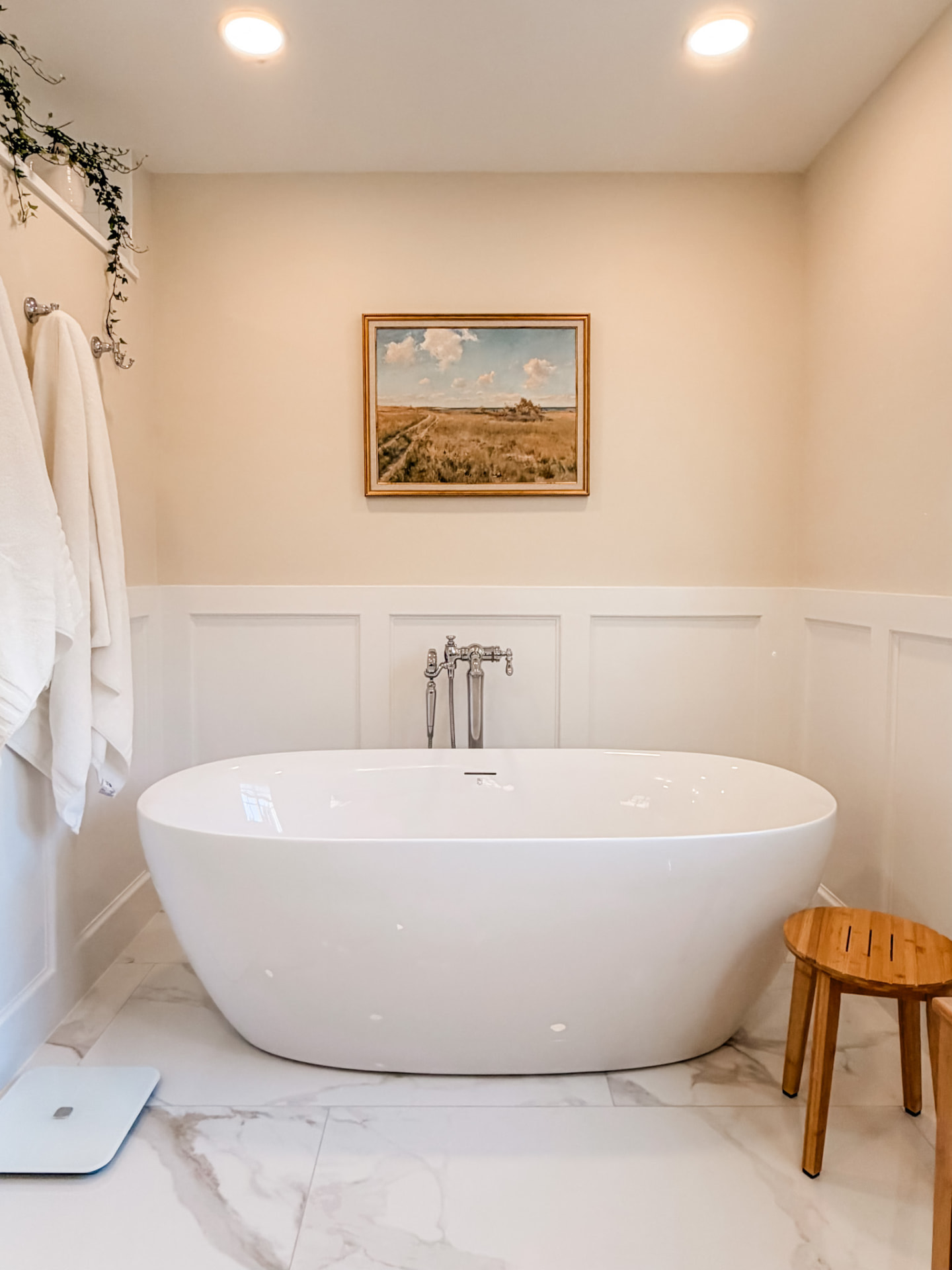Freestanding white soaking tub with floor-mounted polished faucet, paneled walls, and framed landscape artwork