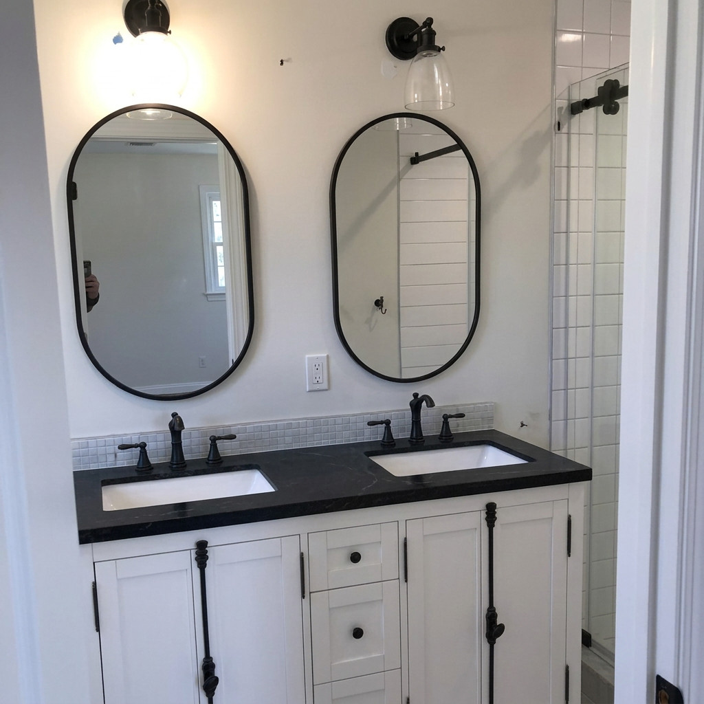 Renovated bathroom with white double vanity, dark stone countertop, matte black faucets, and two oval mirrors beside a tiled shower